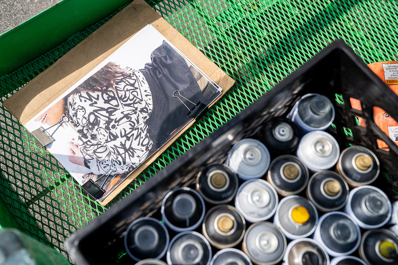 On the floor of a green forklift, reference photos of a girl in a white sweatshirt with a black graffiti design are clipped to cardboard. Next to it is a crate of spray paint.