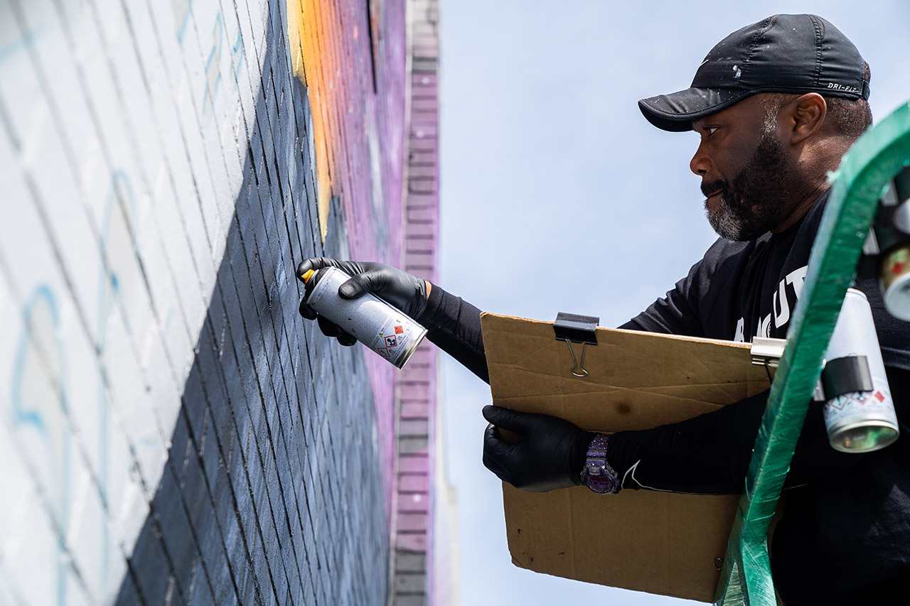 Standing on a green forklift, Rob Gibbs holds his reference photos and sprays paint on the wall, as he finishes his latest mural.