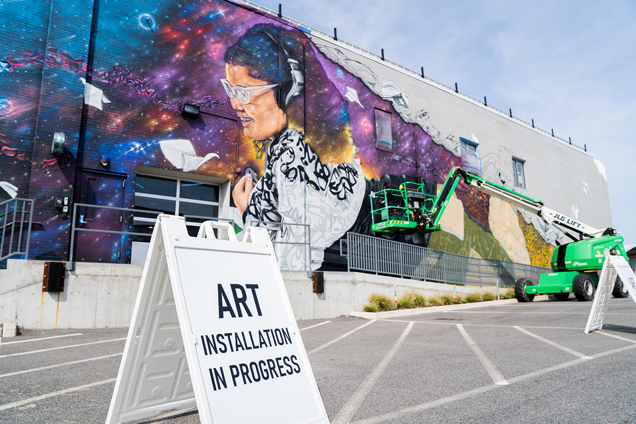 Rob Gibbs stands on a forklift, working on his latest mural at The Record Co. In the foreground, there’s a sign that says, 'Art Installation in progress'