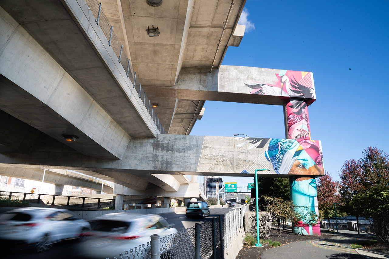 A tall blue and pink mural with a woman’s face and a crane sits on the cement beam holding up I-93, at Underground at Ink Block, as cars fly by on the highway.