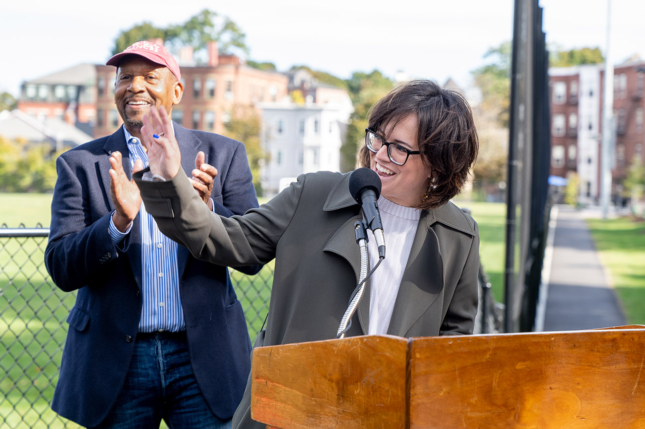 Kara Elliot-Ortega, in an olive green coat and white sweater, stands at a wooden podium and waves at artists in the crowd.