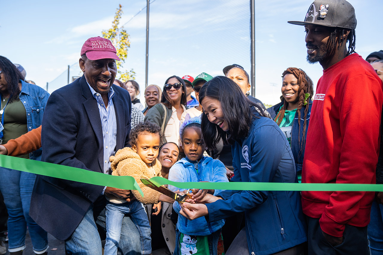 Mayor Michelle Wu, surrounded by smiling community members and children, cut a green ribbon with a pair of golden scissors.