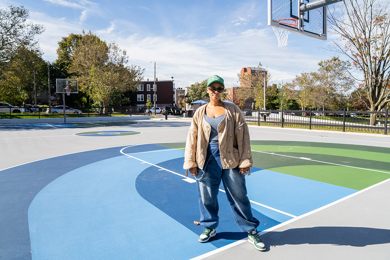Gina Latham, wearing a tan jacket, jeans, a green cap and dark sunglasses, stands on a green and blue basketball court at Malcolm X Park.