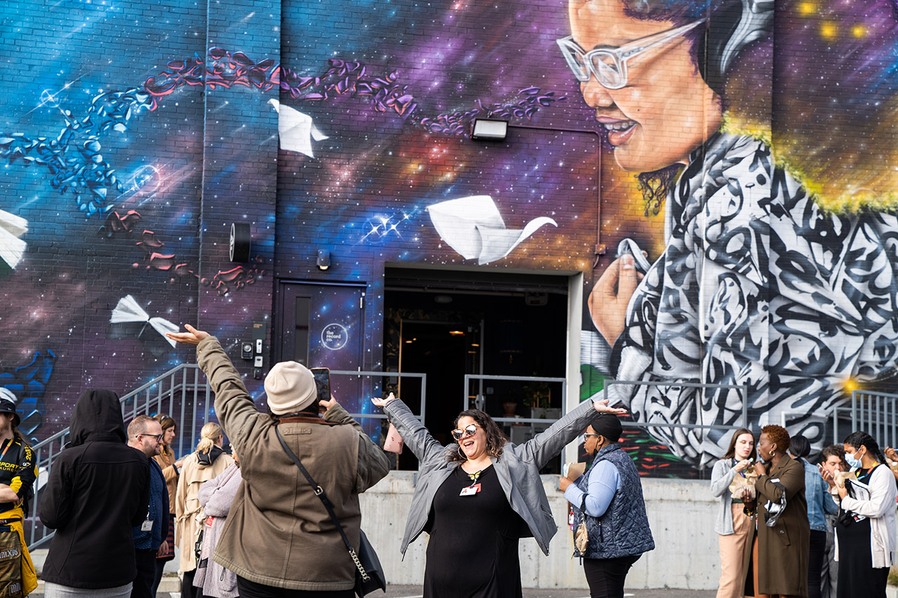 With a crowd of people around her, a woman in a gray jacket and sunglasses holds her arms up in the air and smiles as her photo is being taken in front of Rob Gibbs' new mural at The Record Co.