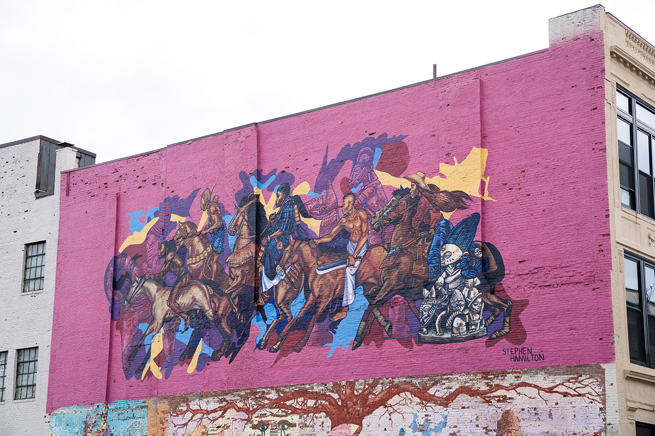 Between beige walls, this mural by Stephen Hamilton shows several people of color riding horses on a pink background.
