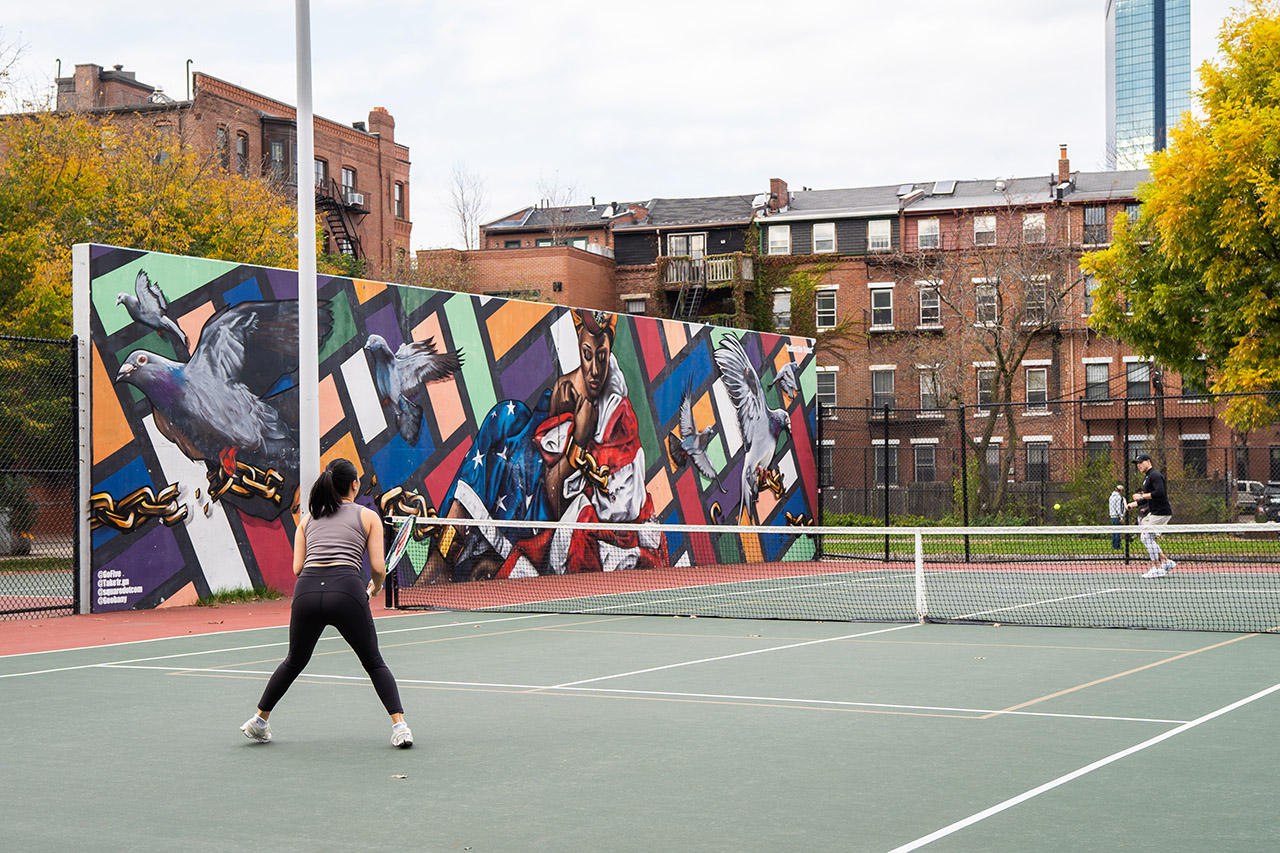 Brett Dietzold, in a black sweatshirt and black cap, hits the tennis ball, as he plays with girlfriend, Sarah Chau, in a gray tank and black leggings. In the background of the court is a colorful mural by GoFive, featuring a woman wrapped in an American flag surrounded by eagles.