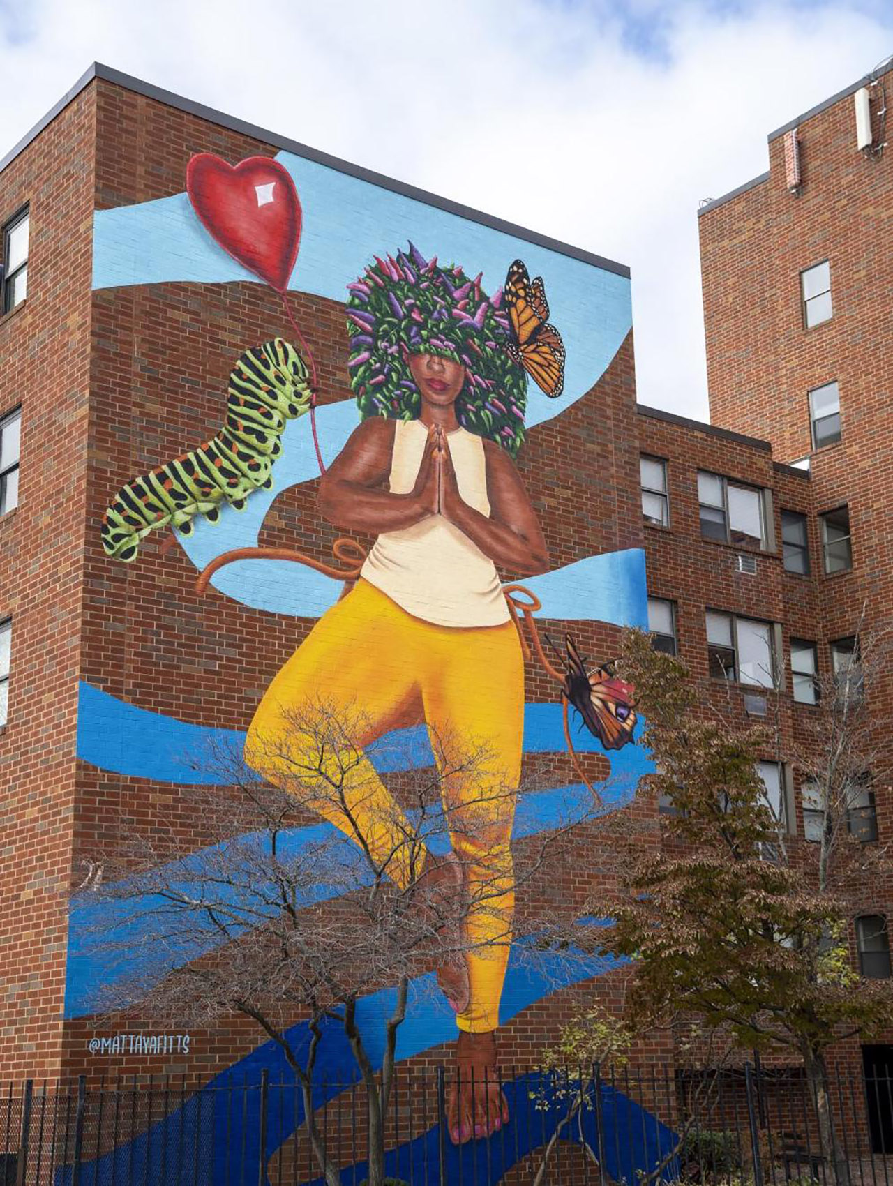 A tall mural sits on a brick wall of an apartment complex, featuring a woman in a one-legged prayer pose who has two monarch butterflies and a caterpillar surrounding her.