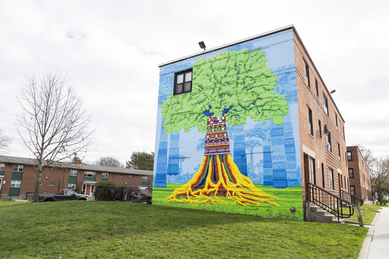 This mural, located on the side of an apartment complex, shows a large tall tree with colorful roots based on the pattern of cultural flags.