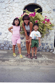 A family of three, including two young children, pose for a photo outside, in front of a wall with flowers.